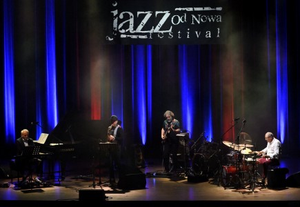 Krzesimir Dębski w otoczeniu innych grających muzyków na scenie. U góry napis: Jazz Od Nowa Festiwal.. Kliknij, aby powiększyć zdjęcie.